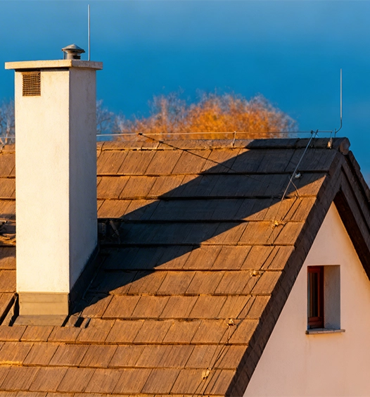 A house roof with brown shingles, a white chimney, a small window, metal rods for lightning protection, and wires, photographed in warm sunlight against a blue sky and autumn trees.