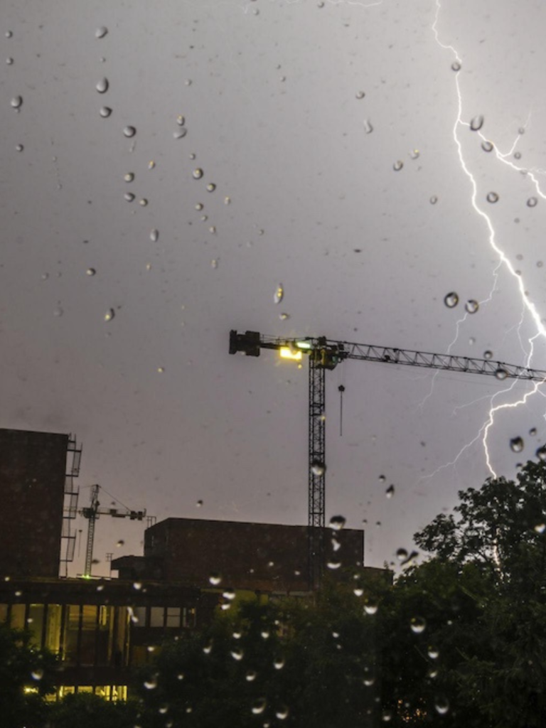 Raindrops on a window with a construction crane, buildings, and a bolt of lightning in the sky during a thunderstorm, highlighting the importance of lightning protection for urban structures.