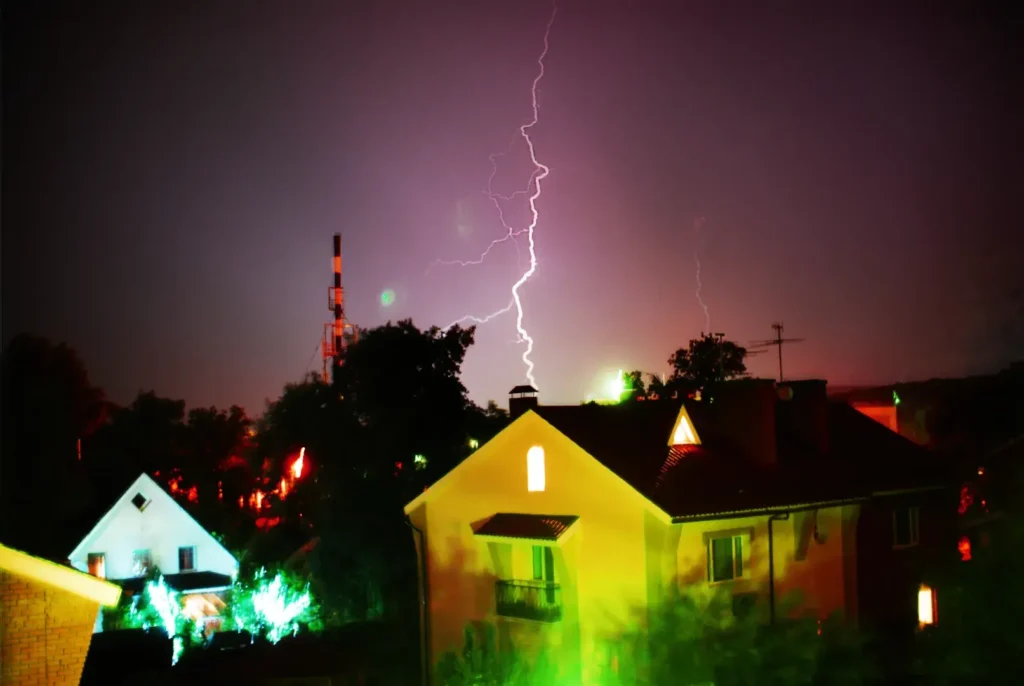 Lightning strikes in the night sky above houses and trees in a residential area, with a communication tower visible in the background—highlighting the importance of lightning protection such as installing a lightning rod.