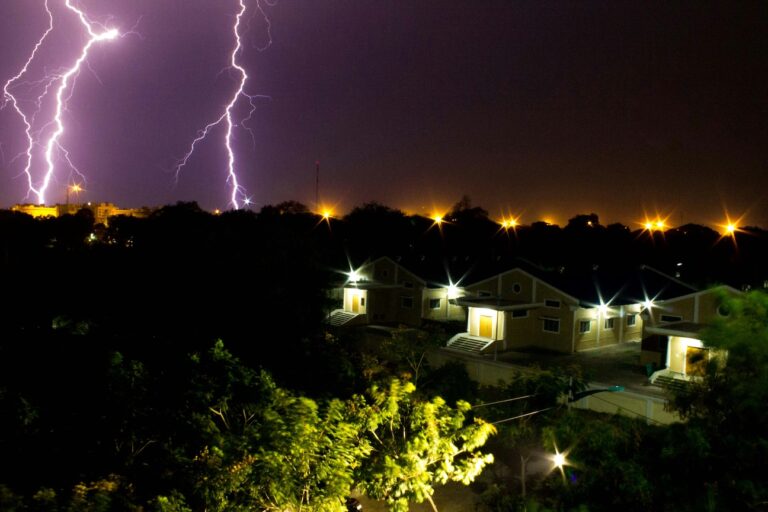 Multiple lightning bolts illuminate a dark night sky above houses and trees, with streetlights and house lights visible—a striking reminder of the importance to install lightning rod systems for reliable lightning protection.