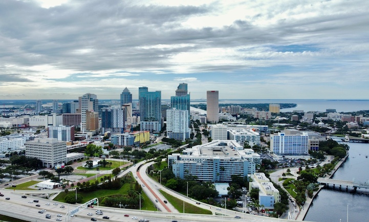 Aerial view of a city skyline with tall buildings, roads, and waterfront under a cloudy sky, highlighting the importance to install lightning rod systems for effective lightning protection.