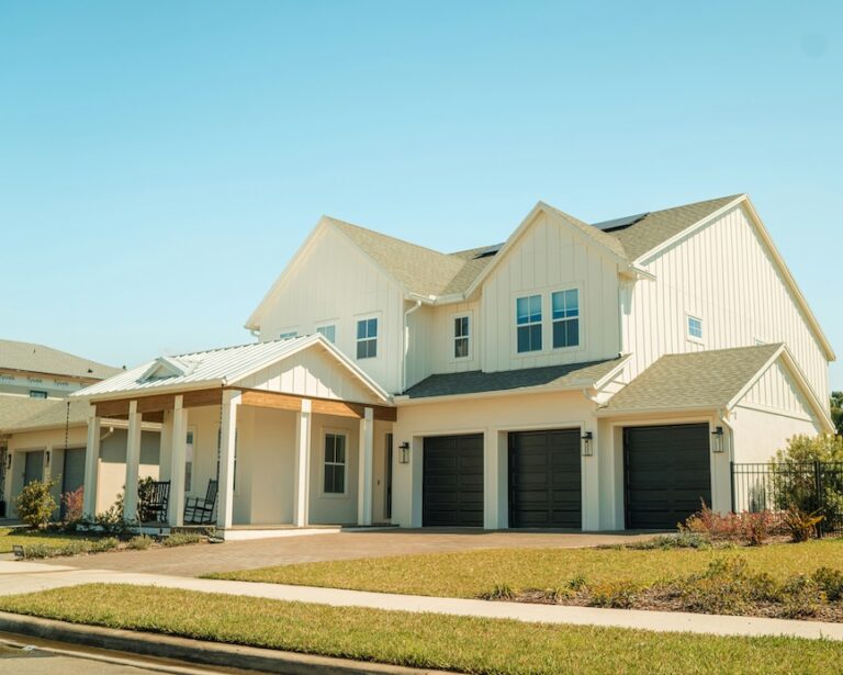 Two-story modern suburban house with white siding, three dark garage doors, a covered front porch, and a well-maintained lawn under a clear blue sky; install lightning rod for added lightning protection.