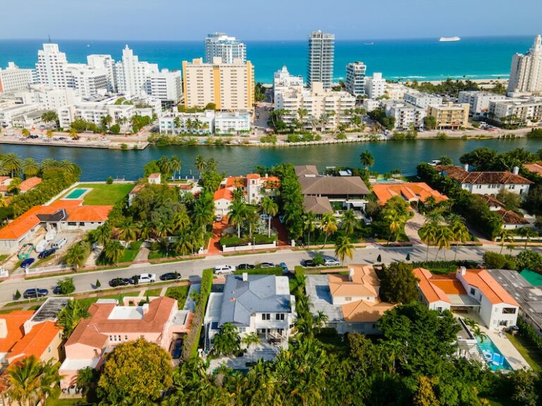 Aerial view of a coastal city with residential houses equipped with lightning protection, palm trees, a canal, high-rise buildings, and the ocean in the background under a clear sky.