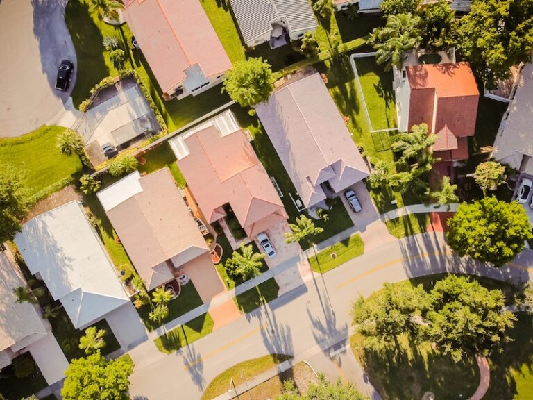 Aerial view of a suburban neighborhood with rows of houses, green lawns, driveways, and tree-lined streets on a sunny day—ideal for those considering lightning protection or planning to install a lightning rod.