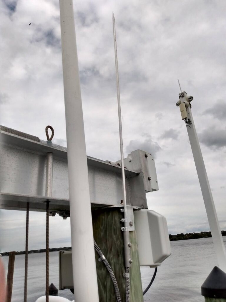 Metal lightning rod and equipment for lightning protection mounted on a wooden pier post by the water, with a cloudy sky in the background.