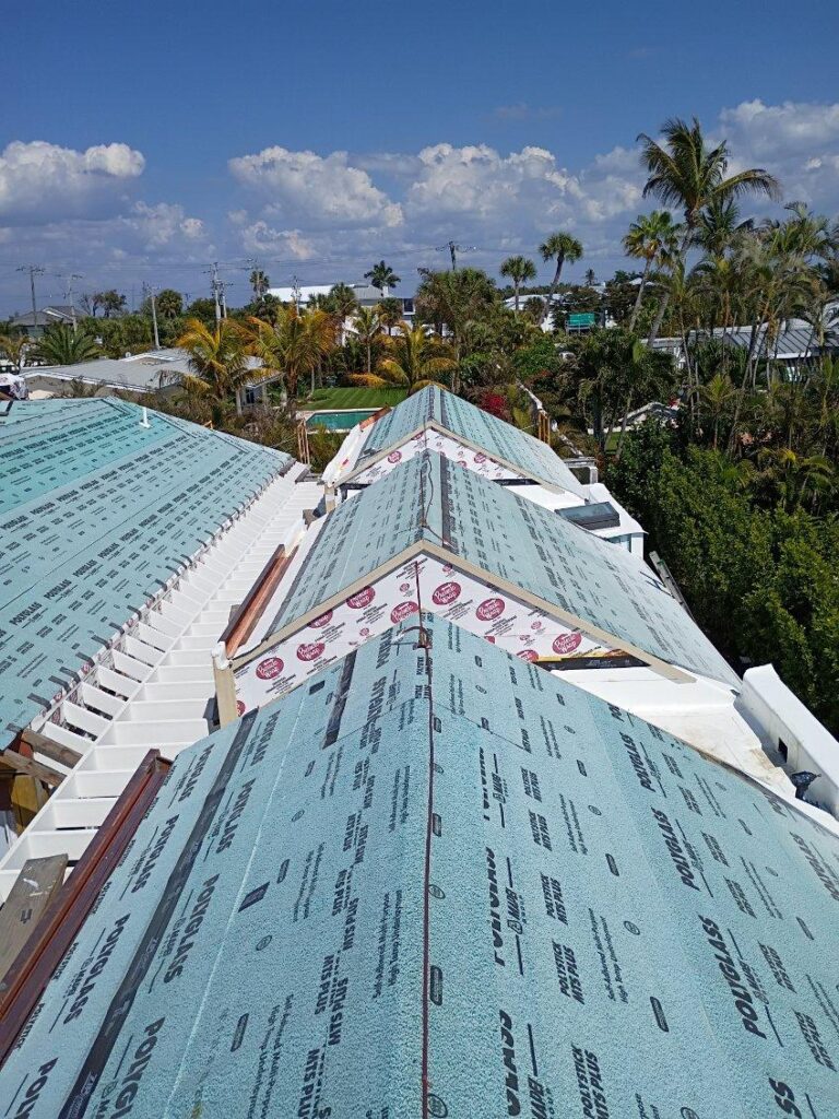 Rooftop under construction with blue synthetic underlayment, labeled tape, and lightning rod installed for lightning protection, surrounded by palm trees and a partly cloudy sky.
