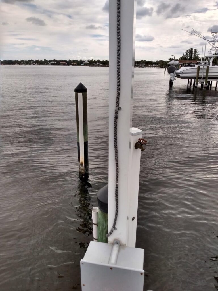A metal post with a rope attached stands on a dock by the water, possibly serving as a lightning rod for added lightning protection, with pilings and boats visible in the background under a cloudy sky.