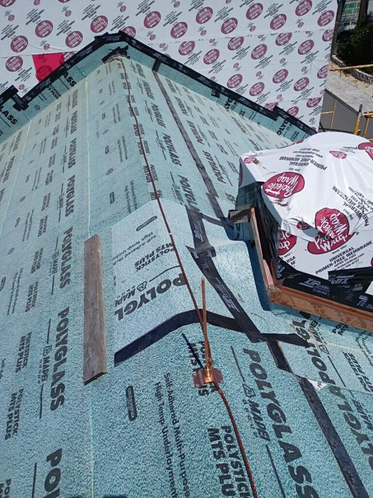 Roof under construction with synthetic underlayment labeled "POLYGLASS," partially installed insulation material, and a bundle of materials near the ridge; ideal stage to install lightning rod for added lightning protection.